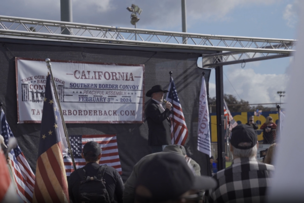 The image shows a speaker on stage in front of a "Take Our Border Back" sign